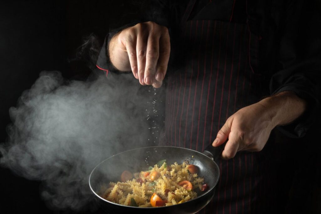 Chef temperando prato de massa com vegetais em meio a vapor durante o reality