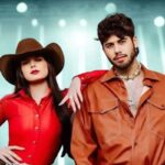 Ana Castela e Zé Felipe posando juntos em figurino country vermelho e laranja com iluminação de palco ao fundo